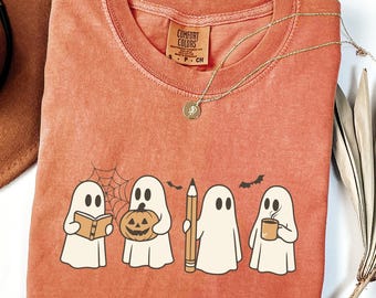 May include: A burnt orange crewneck t-shirt featuring a graphic of four white ghosts. One ghost reads a book, one holds a jack-o'-lantern, one holds a pencil, and one holds a coffee mug. A spiderweb and two small bats are printed above the ghosts.