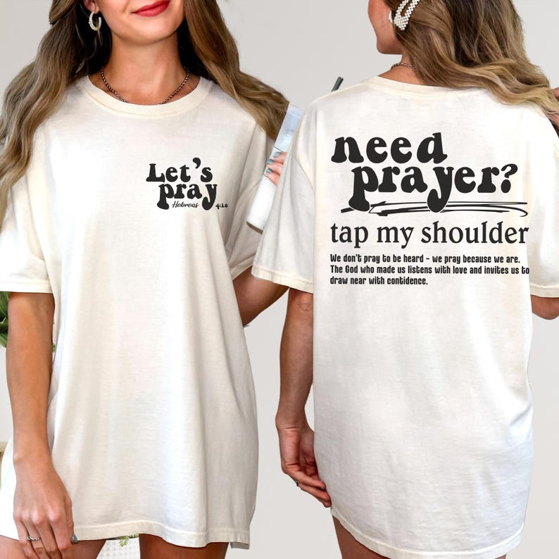 May include: Cream-colored t-shirt with black text. The front reads "Let's Pray" with a Bible verse reference. The back says "Need Prayer? Tap my shoulder" with a supporting quote. A casual, faith-based apparel item.