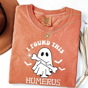 May include: A burnt orange crew neck t-shirt featuring a white graphic of a ghost holding a bone. The text reads I FOUND THIS HUMERUS. The design includes a spider web and three small bats. A gold chain necklace rests on the collar area.