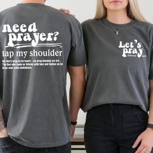 May include: Two gray t-shirts with white text. One shirt says "Need prayer? Tap my shoulder" with a Bible verse. The other says "Let's pray" with a Bible verse. Both shirts are short-sleeved.