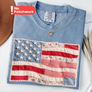 May include: A blue crew neck t-shirt with a Comfort Colors label, featuring a rectangular American flag graphic made of stitched fabric patches in red, white, and blue floral and gingham patterns, with embroidered white stars on a blue field.