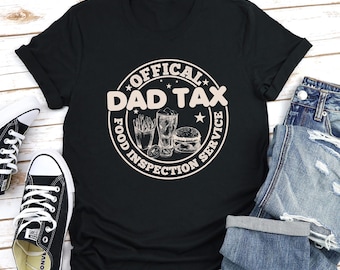 May include: A black short-sleeved t-shirt featuring a circular graphic in light beige. The text reads "OFFICAL DAD TAX" and "FOOD INSPECTION SERVICE" surrounding illustrations of a container of fries, a glass of soda, and a hamburger.