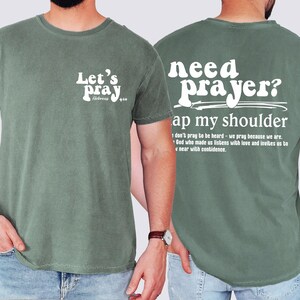 May include: Sage green t-shirt with white text. The front reads "Let's pray" with "Hebrews 4:16" below. The back says "need prayer? tap my shoulder" with a quote below. A casual, comfortable tee.