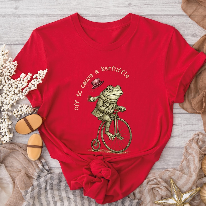 May include: A red t-shirt with a quirky illustration of a frog on a penny-farthing bicycle. The frog is dressed in a jacket, scarf, and hat. The text on the shirt says "off to cause a kerfuffle."