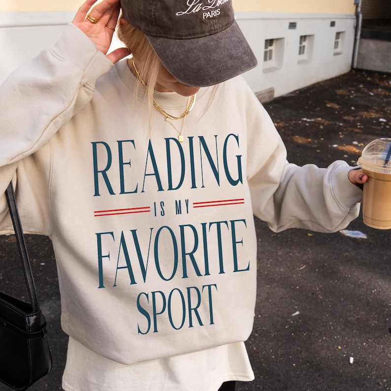 May include: Beige sweatshirt with the text "READING IS MY FAVORITE SPORT" in navy blue. The sweatshirt has a crew neck and long sleeves. A brown baseball cap with white text is worn on the head. A black purse is on the left shoulder.