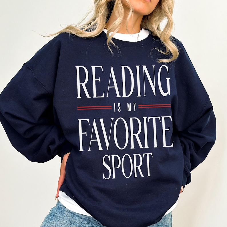 May include: Navy blue sweatshirt with the text "READING IS MY FAVORITE SPORT" in white. The text is separated by two red lines. The sweatshirt is a crew neck style.