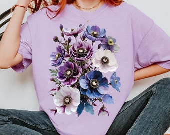May include: A light purple crewneck t-shirt featuring a large, central graphic print of a bouquet of flowers in shades of deep purple, violet, blue, and white. The floral design includes various blooming petals and green leaves arranged in a cluster.