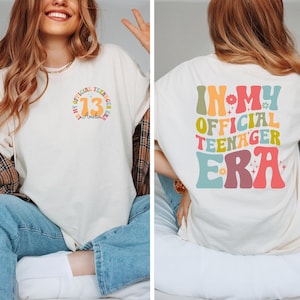 May include: An off-white t-shirt featuring a small circular graphic on the front left chest with the number 13 and the text In My Official Teenager Era. The back displays the large colorful text In My Official Teenager Era with floral accents.