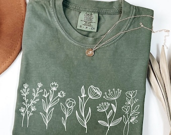 May include: An olive green short-sleeve t-shirt featuring a white line art graphic of various wildflowers across the bottom. The inner neck label reads Comfort Colors, 100% cotton, made in Honduras, size S-P-CH. A thin gold necklace rests on the shirt.
