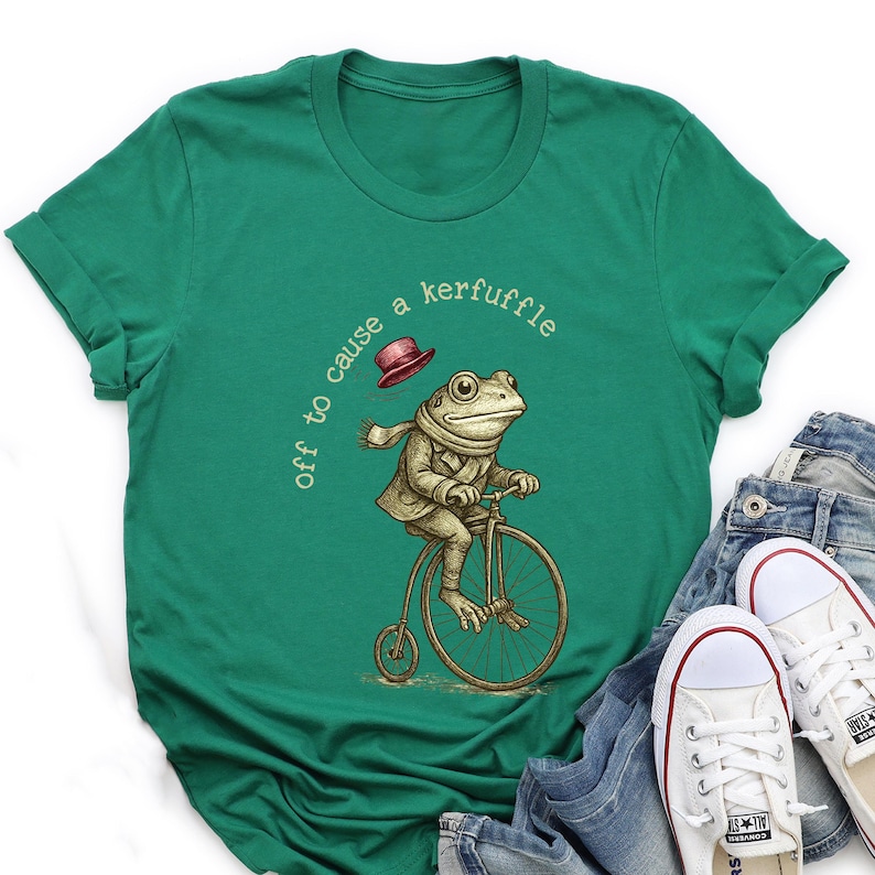 May include: Green t-shirt with a quirky illustration of a frog on a penny-farthing bicycle, wearing a scarf and jacket, and a floating hat. The text "off to cause a kerfuffle" is printed on the shirt.