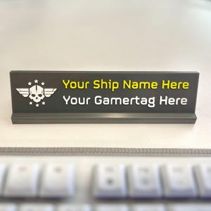 May include: A dark grey desk nameplate featuring a white skull emblem with wings and stars on the left. The text "Your Ship Name Here" is printed in yellow, and "Your Gamertag Here" is printed in white on the front of the rectangular sign.
