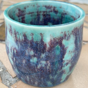 May include: A handmade ceramic cup with a turquoise and deep purple glaze. The cup has a rounded shape and a smooth, glossy finish. The colors blend together, creating a unique and artistic look. This cup is perfect for coffee, tea, or other beverages.
