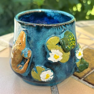 May include: A ceramic mug with a deep blue glaze featuring three-dimensional clay appliques of a brown fish, a green frog with yellow spots, and white water lilies with yellow centers on green lily pads arranged around the exterior surface.
