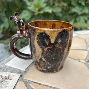 May include: A ceramic mug with a glossy brown and tan glaze featuring a hand-painted African wild dog on the front. The handle is topped with a small, three-dimensional sculpted African wild dog figure with black, brown, and white markings.