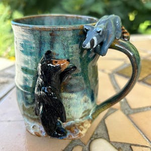May include: A ceramic mug with a glossy teal and blue glaze featuring a three-dimensional black bear climbing the side and a grey raccoon perched on the handle. The mug has a cylindrical shape with a wide opening and a sturdy curved handle.