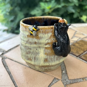 May include: A ceramic mug with a mottled yellow and brown glaze. A small, sculpted black bear is attached to the side, appearing to climb the rim. A tiny, detailed bumblebee with yellow and black stripes is positioned on the mug surface near the bear.
