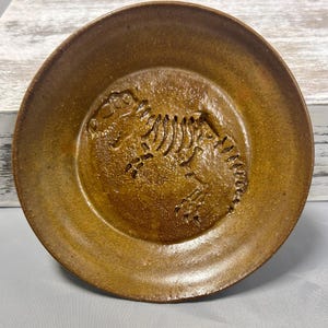 Può includere: Un piatto rotondo in ceramica marrone con un disegno a rilievo di scheletro di dinosauro al centro. Il piatto ha una superficie leggermente irregolare e un tono caldo e terroso, che suggerisce una qualità artigianale. Il bordo è leggermente rialzato.