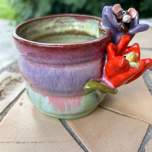 May include: A ceramic mug with a glazed finish in shades of pink, purple, and green. The exterior features a sculpted purple flower with a small mouse figure and a red flower with a small frog figure attached to the side of the rim.
