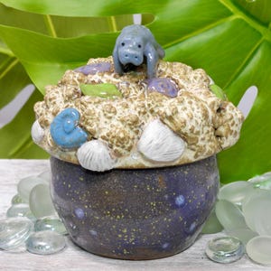 Peut inclure: Un pot en céramique bleu-gris moucheté, surmonté d'un couvercle texturé avec une petite figurine de lamantin bleu. Le couvercle est orné de détails de coquillages et de la vie marine en blanc, bleu et vert.
