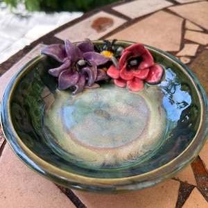 May include: A handcrafted ceramic bowl with a glossy, green and blue glaze. The bowl is adorned with sculpted purple and red flowers, and a yellow and black bee. The bowl is a decorative piece.