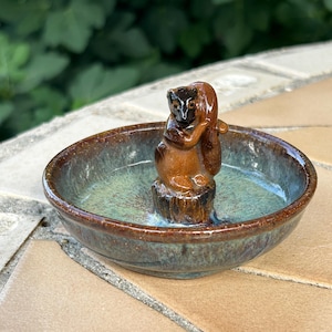 May include: A ceramic squirrel figurine perched on a tree stump in the center of a small, round bowl. The bowl has a brown rim and a teal and brown interior. The figurine is brown with black accents.