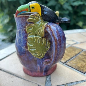 May include: A ceramic mug with a glossy purple and blue glaze featuring a sculpted toucan perched on the handle. The toucan has a black body, yellow neck, and a green, blue, and red beak. Two green monstera leaves are attached to the handle.
