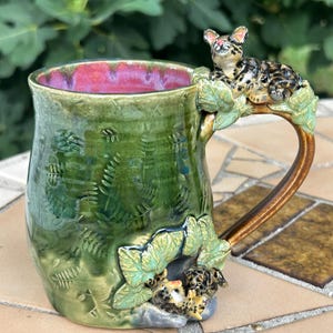 May include: A ceramic mug with a glossy green glaze featuring embossed fern leaf patterns. The handle is shaped like a tree branch with a sculpted leopard resting on top. A small leopard cub is sculpted at the base, and the interior is a vibrant pink.