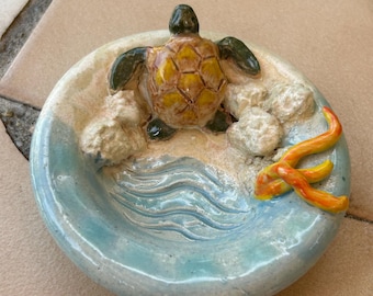 May include: A ceramic ashtray with a beach scene design. The ashtray features a sea turtle, sand, and ocean waves in shades of green, yellow, and blue. An orange starfish adds a pop of colour to the design.