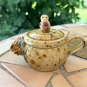 May include: A ceramic teapot with a lid and handle, featuring a small owl figurine on top. The teapot is tan with brown speckles and a decorative floral element on the side. The background is a blurred green.