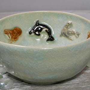 Op de afbeelding: Een lichtblauwe keramische kom met een craquelé glazuur. De kom heeft reliëfsculpturen van zeedieren, waaronder een bruine walrus, een zwart-witte orka, een grijze zeehond en een kleine bruine vis.