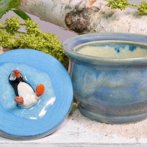 May include: A blue ceramic container with a matching lid. The lid features a raised, detailed puffin design in black, white, and orange. The container has a light blue interior and a darker blue exterior.