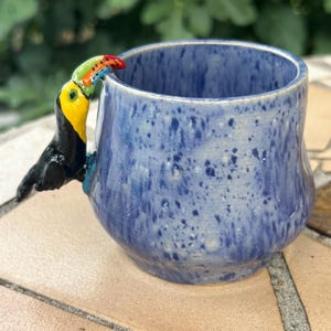 May include: A ceramic mug with a speckled blue glaze and a three-dimensional toucan figurine attached to the side. The toucan features a black body, a bright yellow chest, and a multi-colored beak with red, orange, green, and blue accents.
