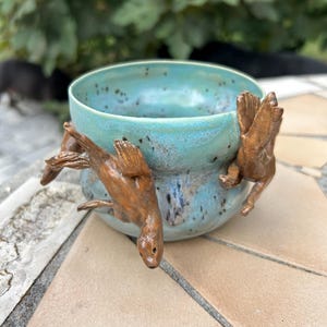 May include: A round ceramic bowl with a glossy turquoise glaze featuring dark speckles. Two sculpted brown figures resembling seals or otters are attached to the exterior, appearing to climb or swim along the curved surface of the vessel.