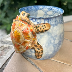 May include: A ceramic mug with a blue and white pattern and a 3D turtle design. The turtle has a brown head and legs, and an orange, yellow, and brown shell. The mug is a unique piece of art.