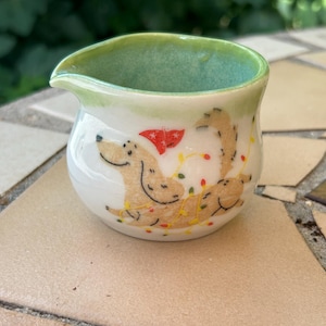 May include: A small, white ceramic creamer with a green interior and rim. The creamer features a whimsical illustration of a dog wearing a Santa hat and Christmas lights. The dog is tan with black outlines.