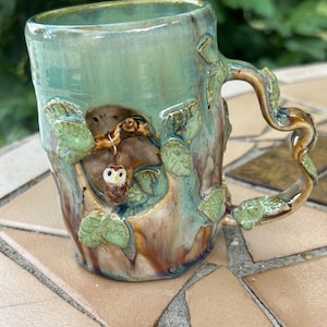 May include: A handcrafted ceramic mug with a nature-inspired design. The mug features a teal glaze, a tree trunk motif, and a sculpted handle resembling a branch. A small owl figurine is nestled within the tree. Green leaves are sculpted around the mug.