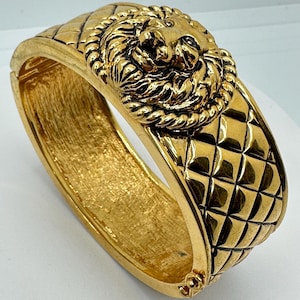 May include: A gold-toned bracelet with a lion's head centerpiece. The bracelet features a quilted pattern on the band and a detailed lion's head with a rope-like border. The clasp is visible on the side. A statement piece of jewelry.