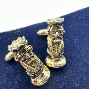 May include: Two gold-colored cufflinks, each shaped like a king chess piece. The king figures have detailed facial features and are wearing crowns. The cufflinks are on a dark blue surface.