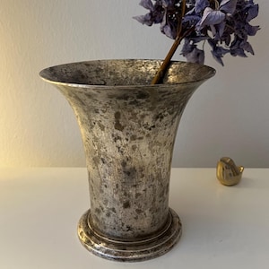 May include: A vintage silver vase with a flared top and a round base, holding a dried purple flower. A small gold bird figurine sits to the right of the vase. The vase has a distressed, aged appearance.