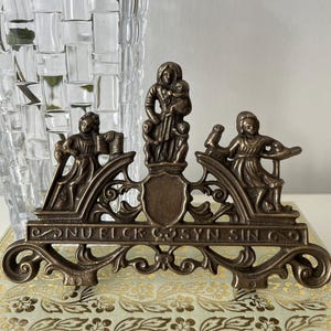 Könnte beinhalten: Ein bronzefarbenes dekoratives Metallstück mit drei Figuren und dem Text "DNU ELICK SYN SIN O". Die zentrale Figur hält ein Kind. Das Stück liegt auf einem Buch mit goldenem Blumenmuster, mit einer durchsichtigen Glasvase im Hintergrund.
