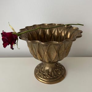 Könnte beinhalten: Eine verzierte, goldfarbene Zierschale mit gewelltem Rand und Sockel. Die Schale hat eine strukturierte Oberfläche und florale Muster. Eine einzelne, tiefrote Rose liegt am Rand der Schale.