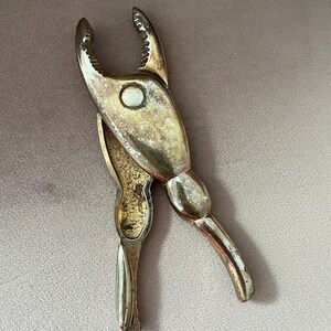 Pode incluir: Um quebra-caranguejo prateado vintage. A ferramenta tem uma extremidade de preensão em forma de garra e uma alça para alavancagem. O metal mostra sinais de desgaste, adicionando ao seu charme antigo. Um utensílio de cozinha clássico, com cerca de 15 cm de comprimento.