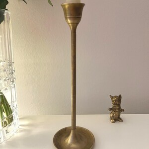 Puede incluir: Un candelabro alto de latón antiguo con una vela blanca. El candelabro tiene una base redonda y un tallo delgado. A la derecha del candelabro hay una pequeña figura de gato de latón. Un jarrón de cristal transparente con rosas rosas en el fondo.