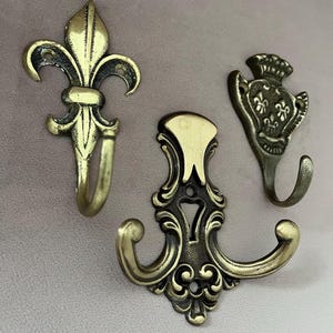 Könnte beinhalten: Drei antike Wandhaken aus Messing mit verzierten Designs. Die Haken umfassen eine Fleur-de-Lis, einen Doppel-Haken und ein Kronen-Motiv. Sie sind auf einer hellrosa Oberfläche montiert, die ihre komplizierten Details und ihre Vintage-Ästhetik hervorhebt.