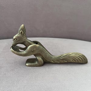 Könnte beinhalten: Ein metallischer, messingfarbener Nussknacker in Form eines Eichhörnchens. Das Eichhörnchen ist in sitzender Position dargestellt, wobei der Schwanz horizontal ausgestreckt ist. Die Vorderpfoten sind erhoben und bilden eine gewölbte Öffnung.