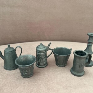Op de afbeelding: Een verzameling miniatuuritems van tin, waaronder een theepot, twee kopjes, een bierpul, een emmer, een klein vat en een kandelaar. De items zijn allemaal donkergrijs van kleur en lijken vintage of antiek te zijn.