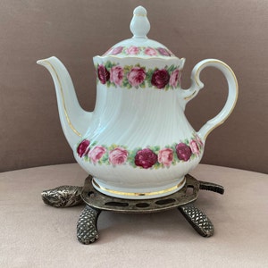 Puede incluir: Una tetera de porcelana blanca con un diseño floral y ribetes dorados se asienta sobre un soporte de metal con forma de tortuga.