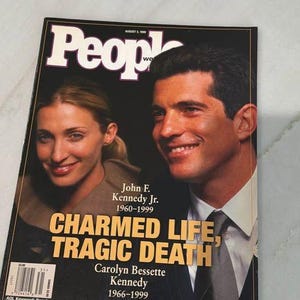 Pode incluir: Uma edição da revista People de 2 de agosto de 1999 com uma fotografia de capa de John F. Kennedy Jr. e Carolyn Bessette Kennedy. A manchete diz Charmed Life, Tragic Death, com seus nomes e anos de nascimento e morte abaixo.