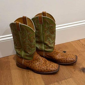 May include: A pair of western-style boots featuring a textured brown leather foot with a distinct quill pattern and a tall olive green leather shaft. The shaft is decorated with intricate orange and green stitching, white piping, and brown pull tabs.