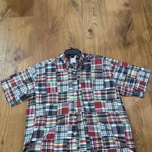 May include: A short-sleeve button-down shirt featuring a patchwork design composed of various multicolored plaid patterns in shades of red, navy blue, white, green, and yellow. The shirt has a classic collar and a front button placket.